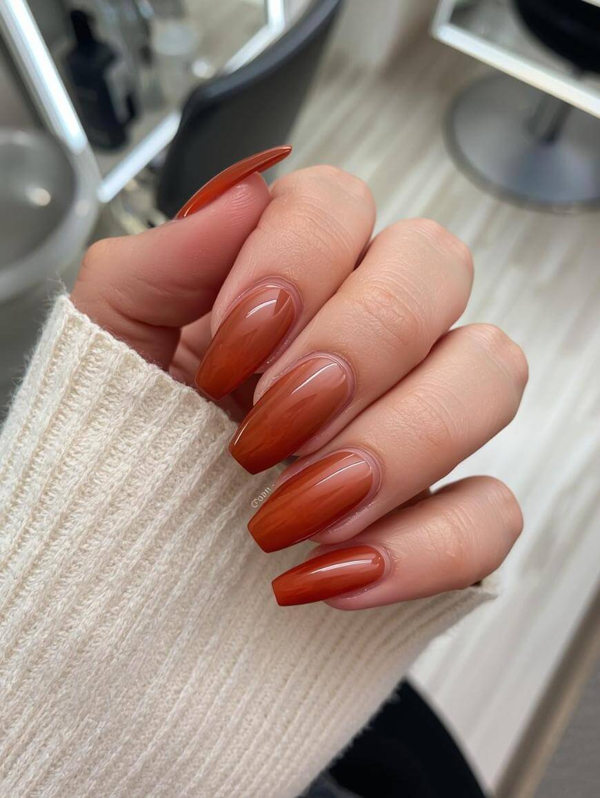 A hand with long, almond-shaped nails painted in a glossy burnt orange shade, resting on a ribbed cream-colored sweater. A salon chair and mirror are visible in the blurred background.
