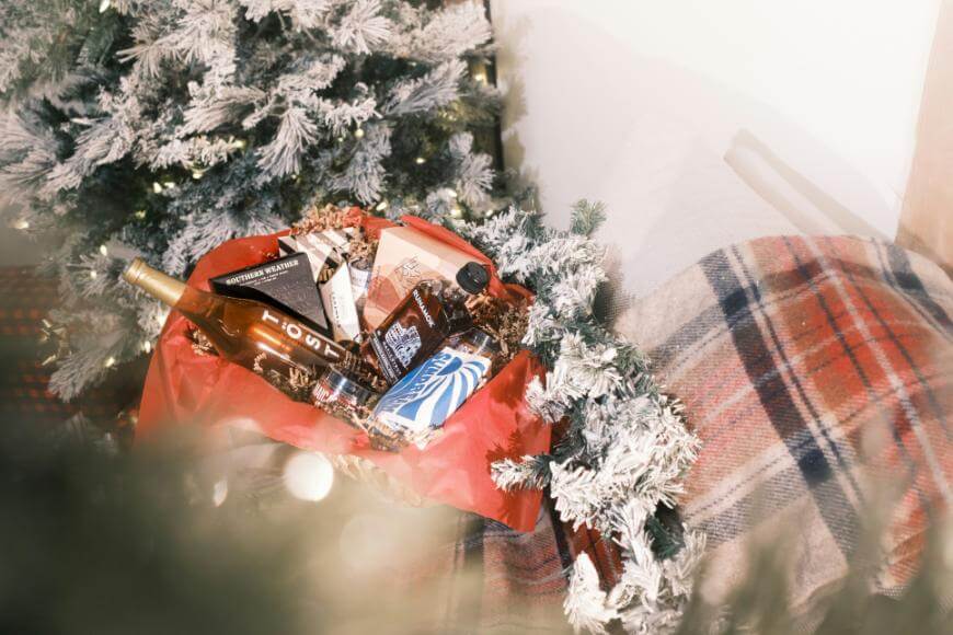 A festive gift basket filled with various treats and a bottle, wrapped in red tissue paper, sits next to a snow-dusted Christmas tree and a plaid blanket.