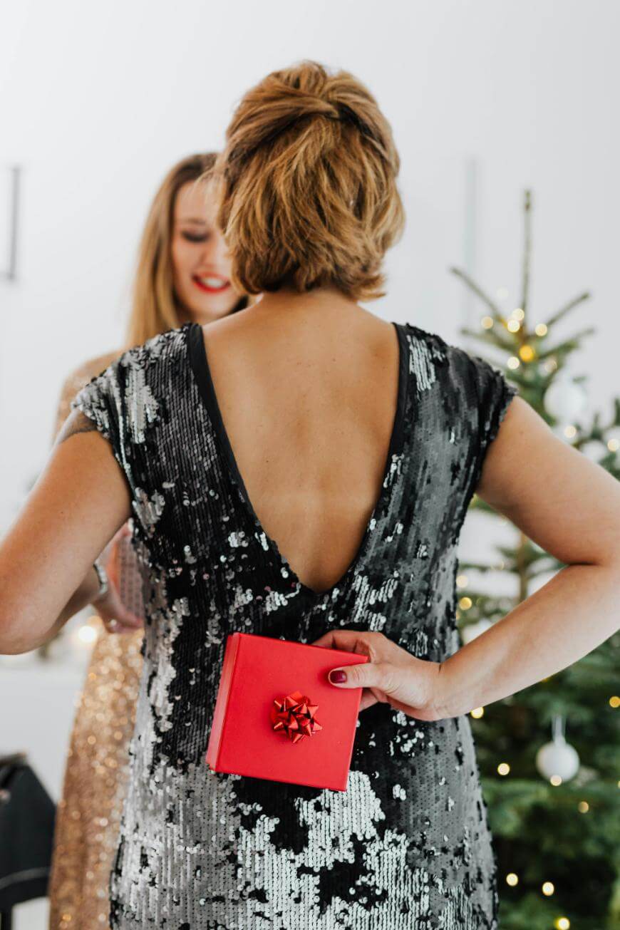 A woman in a sequined dress holds a small red gift with a bow behind her back, standing near a decorated Christmas tree, while another woman in a sparkly dress smiles in the background.