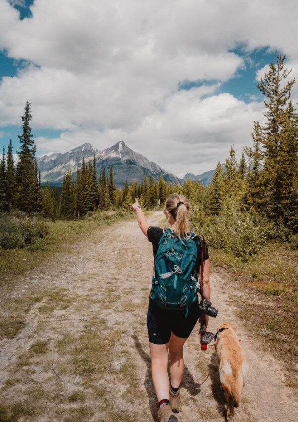 A woman with a large teal backpack walks on a dirt trail through a forest, pointing toward distant mountains, accompanied by a golden retriever dog under a partly cloudy sky.
