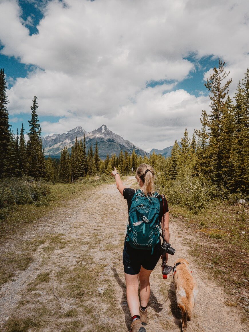 A woman with a backpack and camera walks on a mountain trail with a golden retriever, pointing toward distant snow-capped peaks under a partly cloudy sky, surrounded by pine trees.