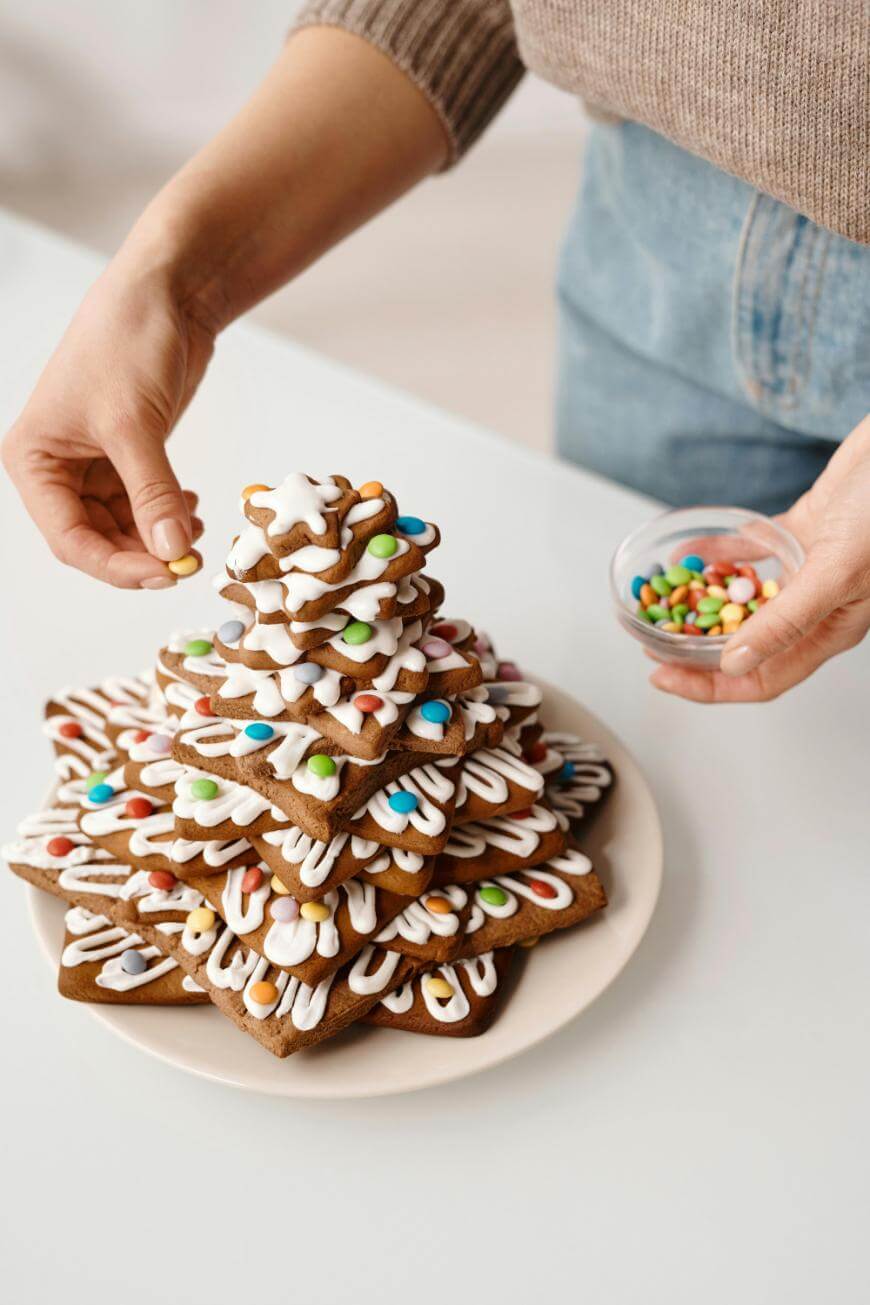 A person decorates a stacked gingerbread cookie tree with colorful candy and white icing. The cookies are star-shaped and arranged by size on a white plate. The person holds a small bowl of candies.