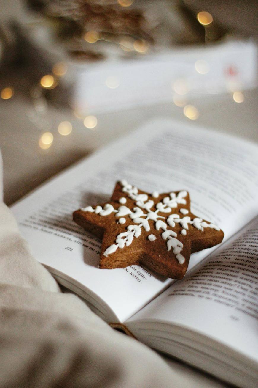 A star-shaped gingerbread cookie with white icing decoration rests on the open pages of a book, with soft, warm lights blurred in the background.