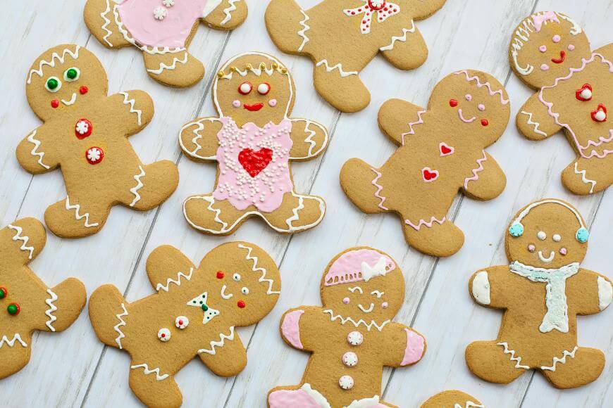 Decorated gingerbread cookies shaped like people are arranged on a white wooden surface. Each cookie is decorated with colorful icing and candies, featuring different patterns, faces, and accessories.