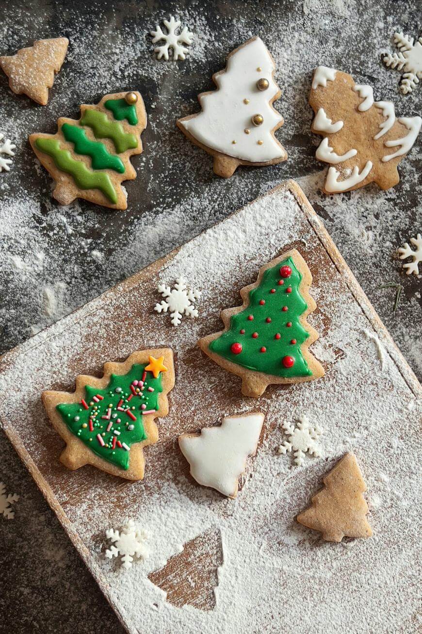 Christmas tree-shaped cookies decorated with green and white icing, some with sprinkles, are arranged on a floured surface and a wooden board, surrounded by small snowflake decorations.