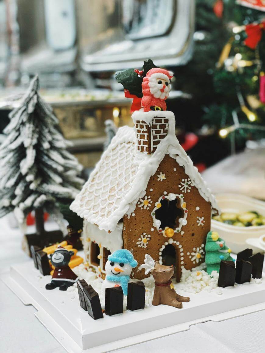 A decorated gingerbread house with white icing, candy decorations, and a Santa figure on the roof. Snowmen and a reindeer are nearby, with a Christmas tree and festive decorations in the background.