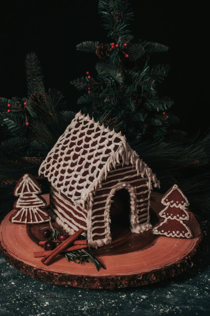 A gingerbread house decorated with white icing sits on a wooden platter, flanked by two gingerbread trees, cinnamon sticks, and festive greenery, with pine branches in the background.