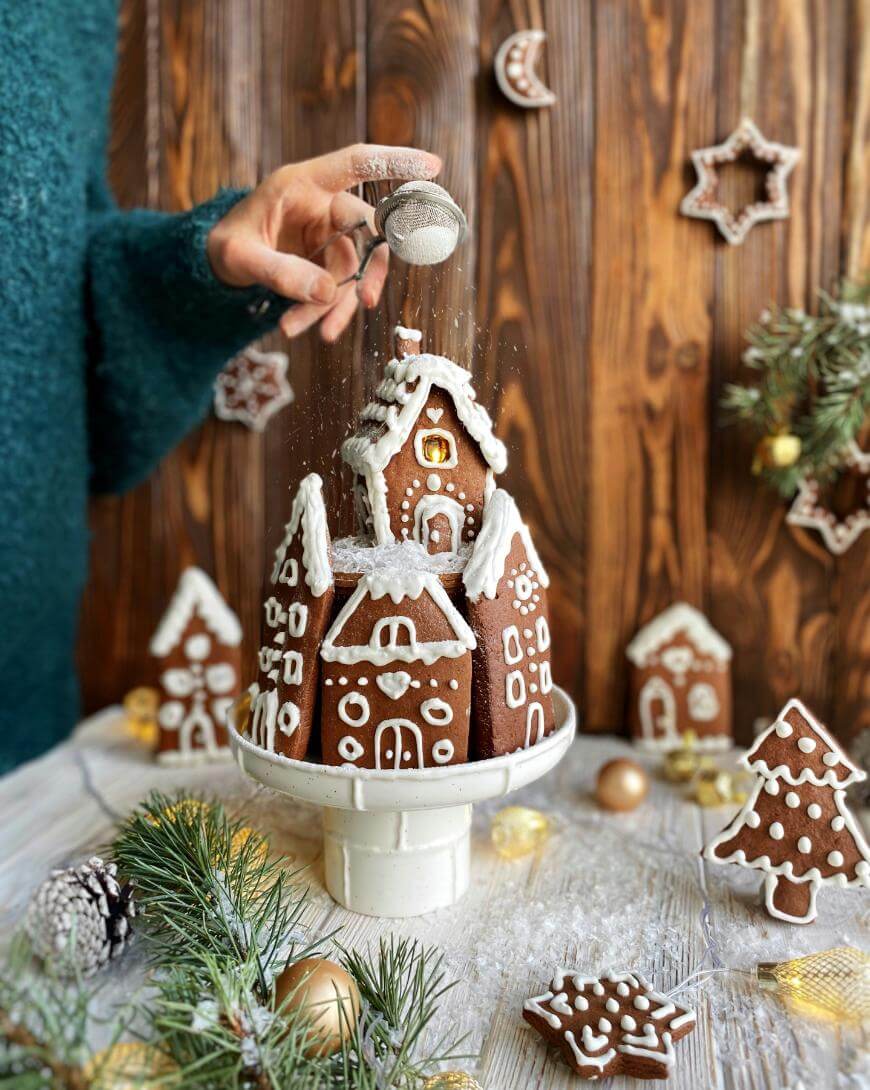 A hand sprinkles powdered sugar onto a decorated gingerbread house on a cake stand, surrounded by gingerbread cookies, pine branches, and holiday decorations on a wooden background.