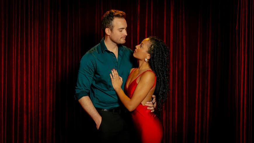 A man in a teal shirt and black pants stands closely with a woman in a sleeveless red dress, who has her hand on his chest. They gaze into each other’s eyes, standing in front of red curtains.