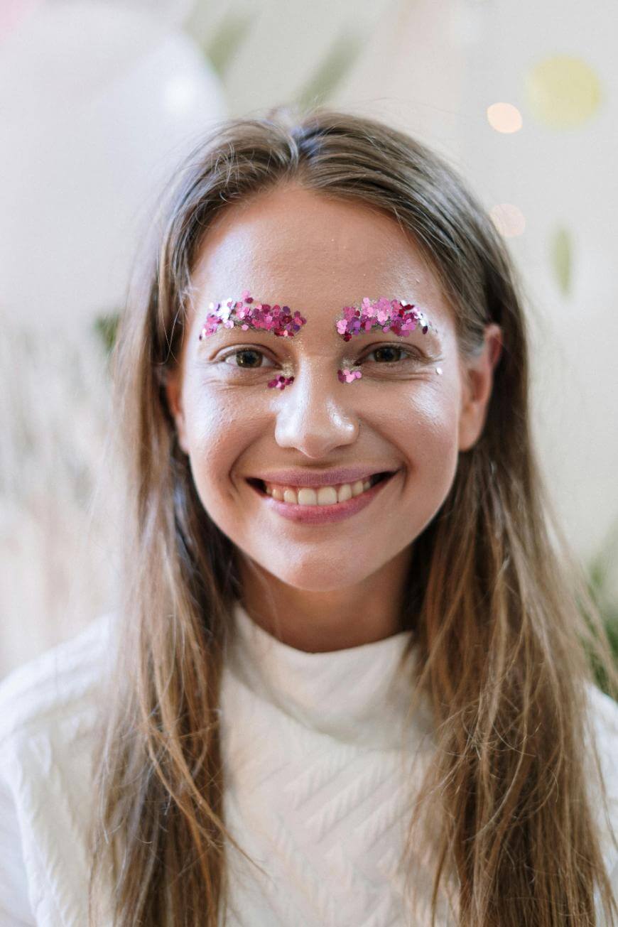 A woman with long brown hair smiles at the camera, wearing a white textured top. She has pink and purple sequins decorating her eyebrows, giving a festive and artistic appearance.
