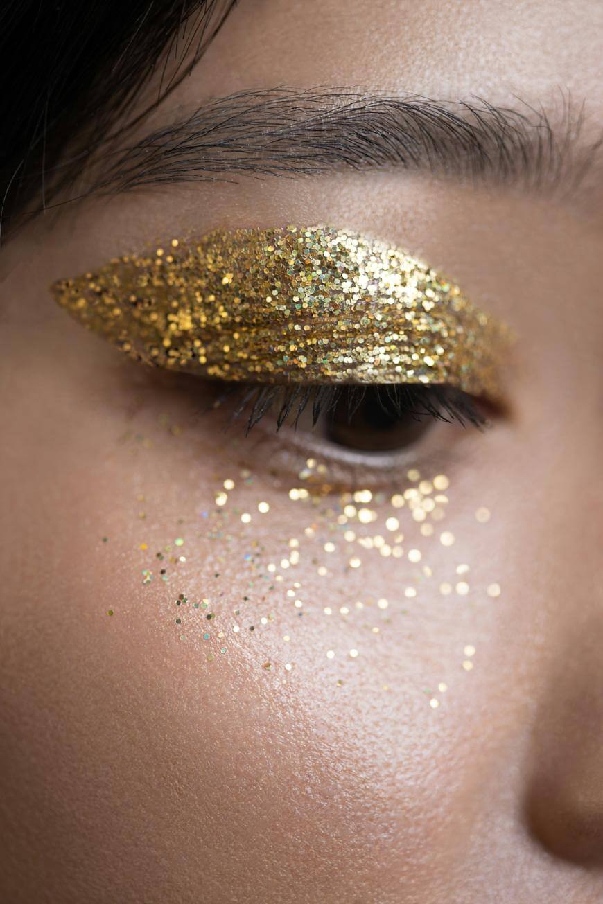 Close-up of a person’s closed eye with bold gold glitter eyeshadow on the eyelid and scattered gold glitter below the eye on smooth, natural skin.