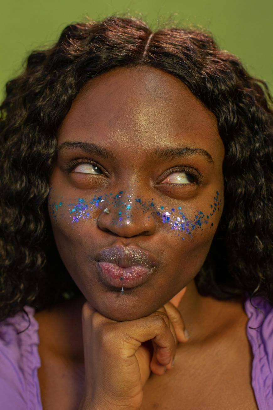A person with curly dark hair, wearing purple clothing, has blue and silver glitter on their cheeks and nose. They are looking up thoughtfully with pursed lips, resting their chin on their hand, against a green background.