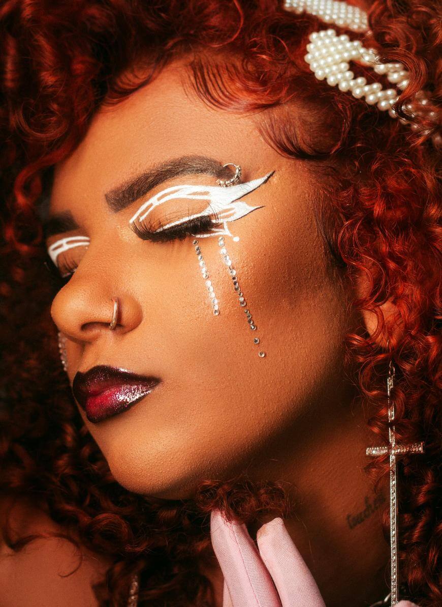 A person with vibrant red curly hair and elaborate white eyeliner, rhinestones under the eyes, dark glossy lipstick, a nose ring, and a pearl hair accessory poses with eyes closed in dramatic makeup.