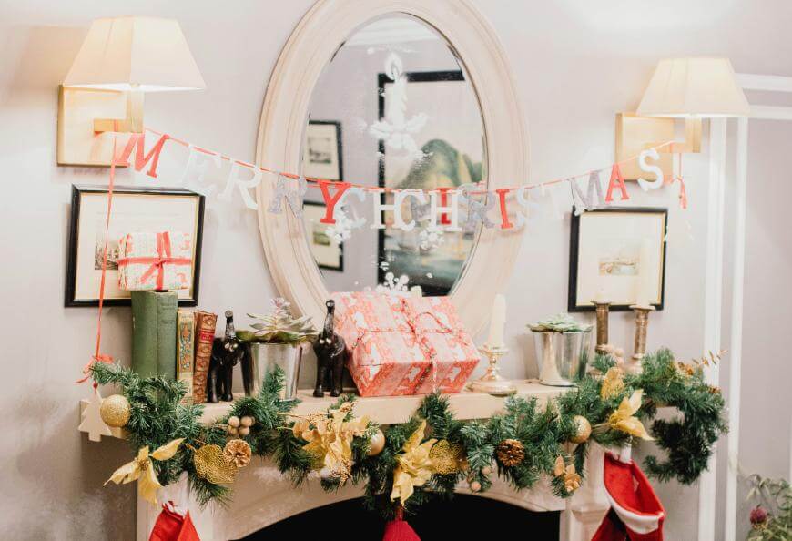 A festive fireplace mantel decorated with greenery, gold ornaments, stockings, wrapped gifts, books, small statues, and a Merry Christmas banner. A large mirror and framed pictures hang above.