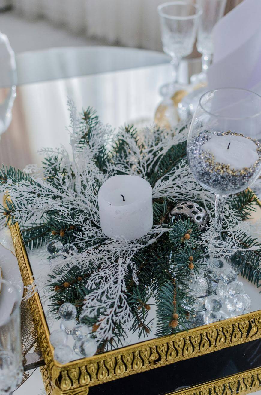 A decorative centerpiece features a white candle surrounded by pine branches, frosted white foliage, glass beads, and a gold-trimmed mirrored tray. A wine glass with silver and gold decorations stands nearby.