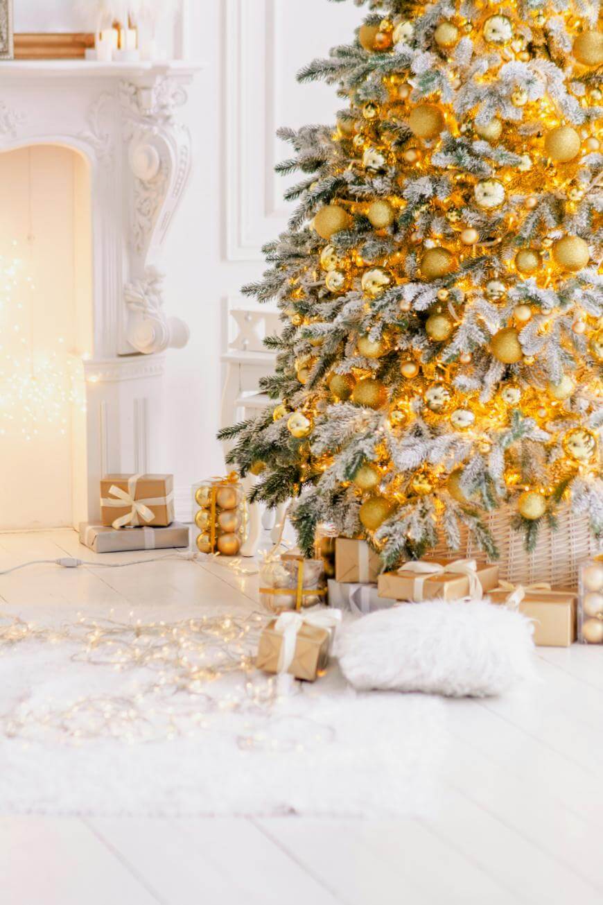 A Christmas tree decorated with gold ornaments and lights stands beside a white fireplace. Wrapped presents in neutral tones are placed under the tree and on a white rug, creating a cozy and festive atmosphere.