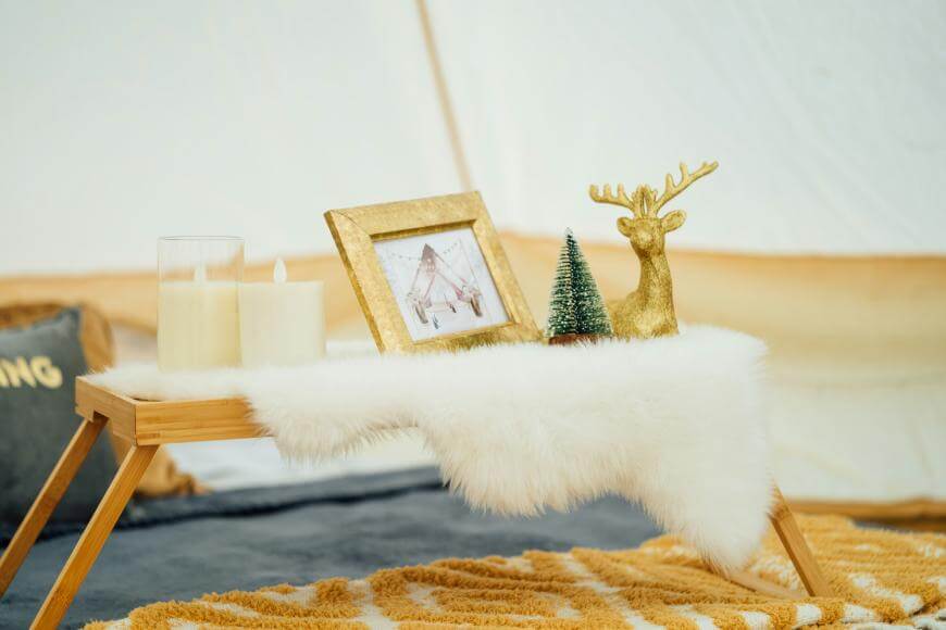 A small wooden tray with a white fur cover holds two pillar candles, a gold-framed photo, a miniature fir tree, and a gold reindeer figurine, set on a cozy rug with soft furnishings in the background.