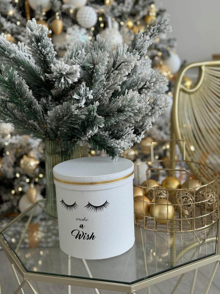A white gift box with eyelash illustrations and the words make a wish sits on a glass table beside a frosted pine branch. A gold wire basket with gold ornaments and a decorated Christmas tree are in the background.