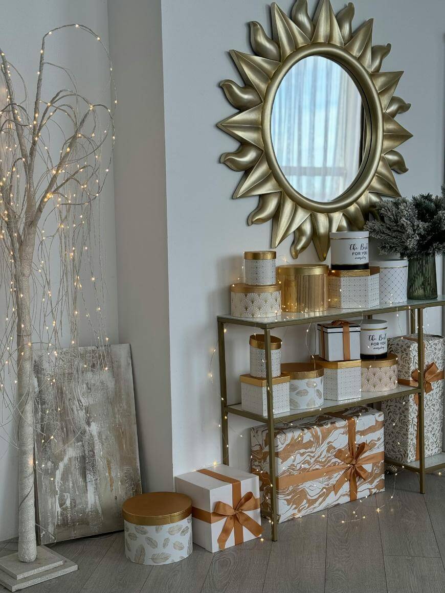 A gold sunburst mirror hangs above gold shelves holding decorative boxes and vases. Wrapped gifts with gold and white patterns sit below. A lit, white branch tree and abstract art lean against the wall.