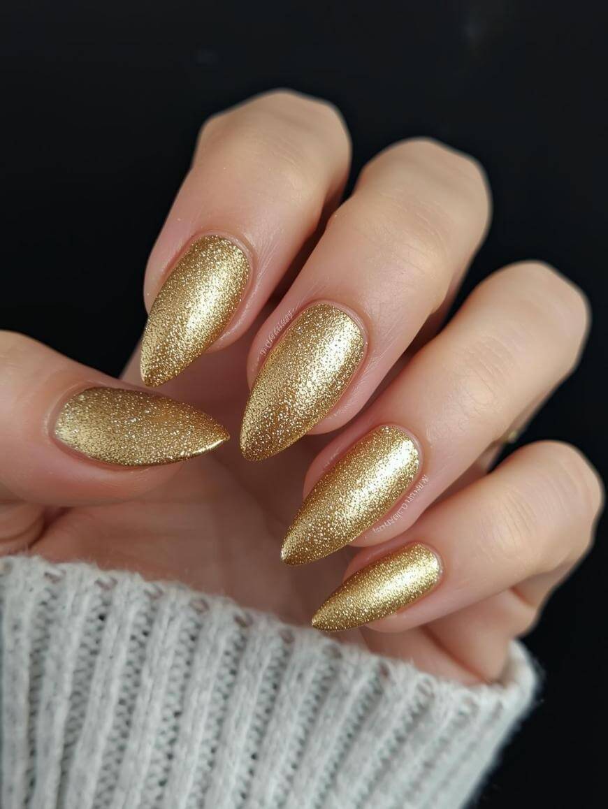 A hand with long, almond-shaped nails painted with sparkly gold nail polish. The person is wearing a light gray, ribbed sweater. The background is black.