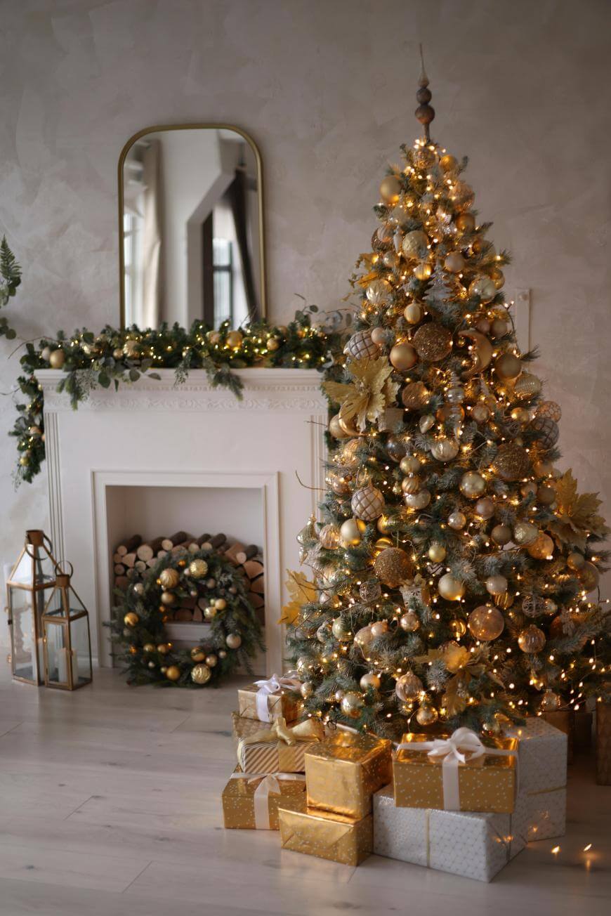 A decorated Christmas tree with gold ornaments and lights stands beside a white fireplace adorned with garland and a wreath. Several wrapped gifts in gold and white paper sit on the light wooden floor.