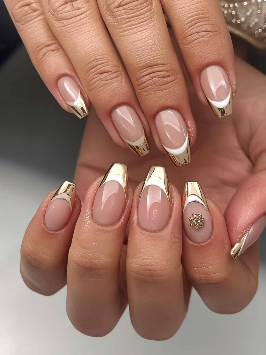 A close-up of manicured hands with nude pink nails, featuring shiny gold French tips and one accent nail adorned with a small gold embellishment.