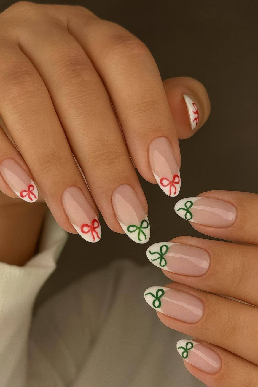 Close-up of hands with almond-shaped nails, featuring a nude base and white tips. Each tip is decorated with a small red or green bow design. The overall style is neat and festive.