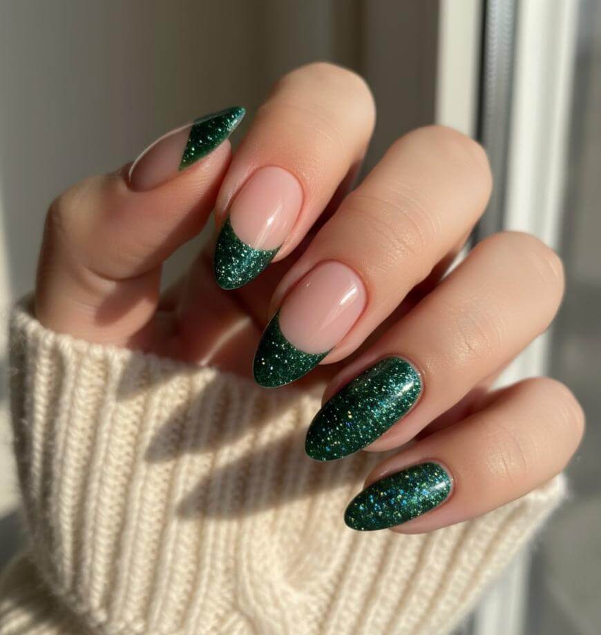 A hand with almond-shaped nails features a green glittery French tip design. The person is wearing a cream-colored ribbed sweater, and light shines softly through a nearby window.