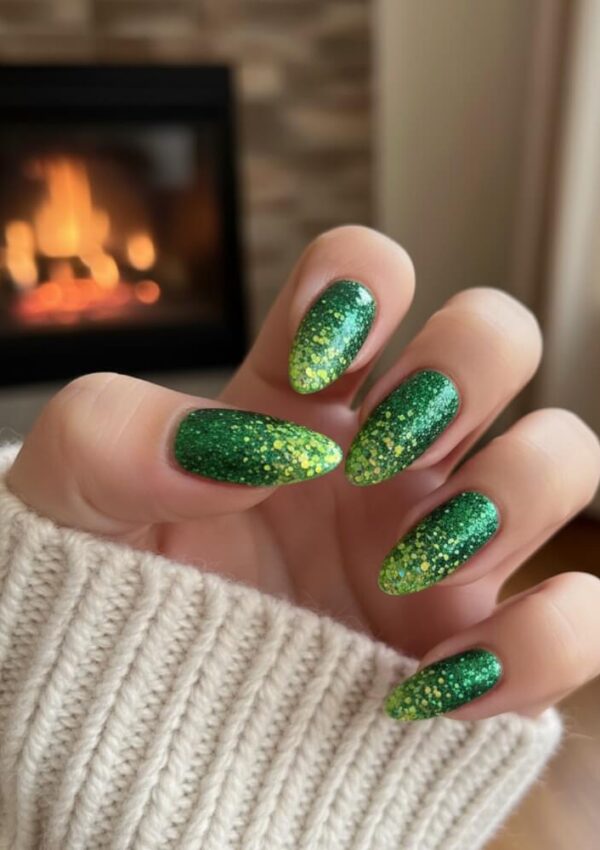 A hand with long, almond-shaped nails painted in sparkly green and yellow ombre glitter polish, shown against a neutral cozy sweater and a blurred background with a lit fireplace.
