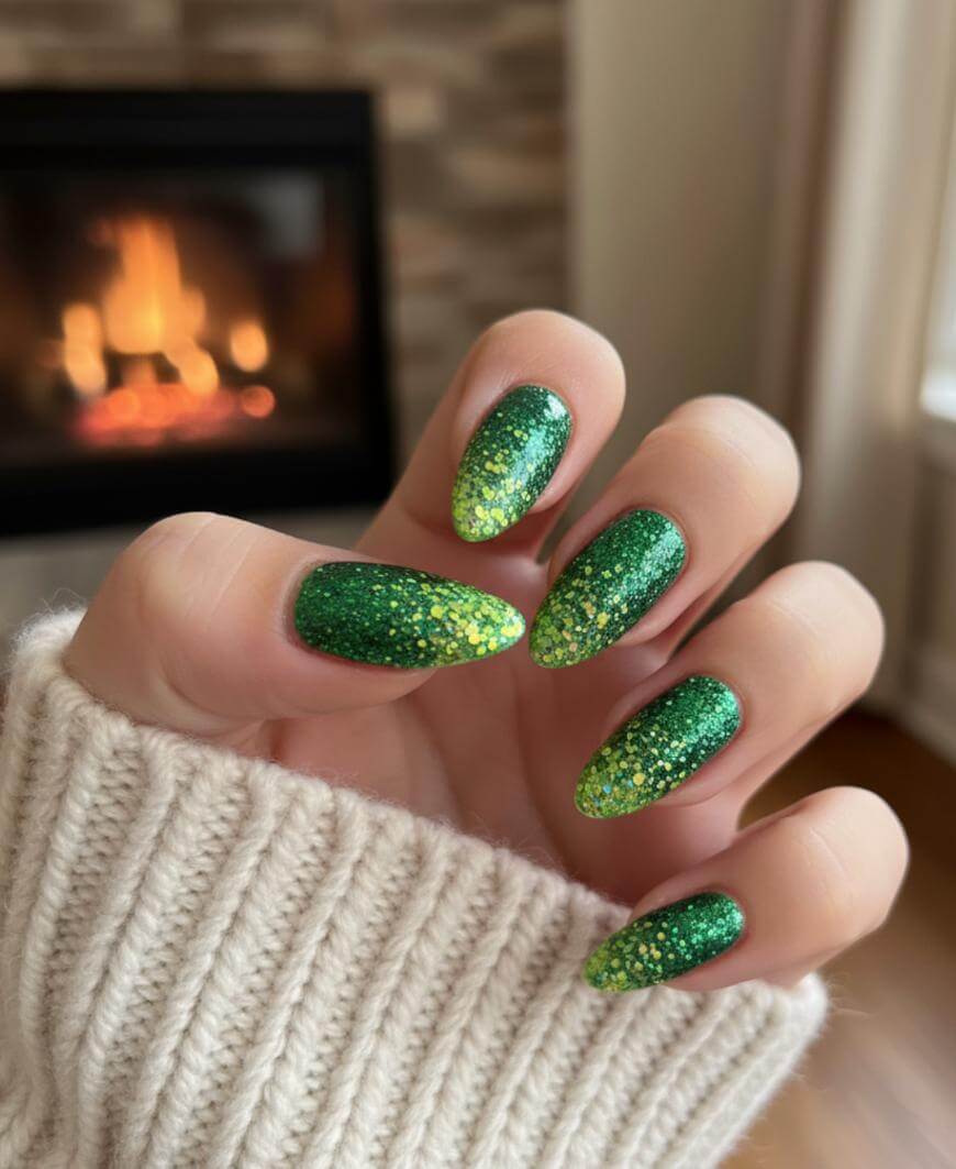 A hand with long, almond-shaped nails painted in green and yellow glitter polish, held up in front of a fireplace. The person is wearing a cream-colored knit sweater.
