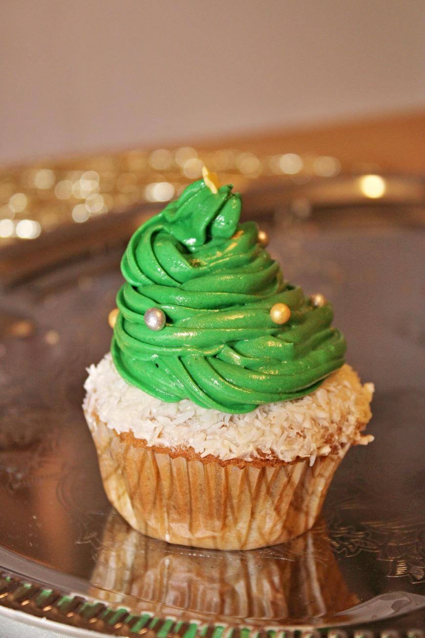 A cupcake decorated with green frosting shaped like a Christmas tree, topped with small gold and silver candy balls, and white coconut flakes around the base, sitting on a silver tray.