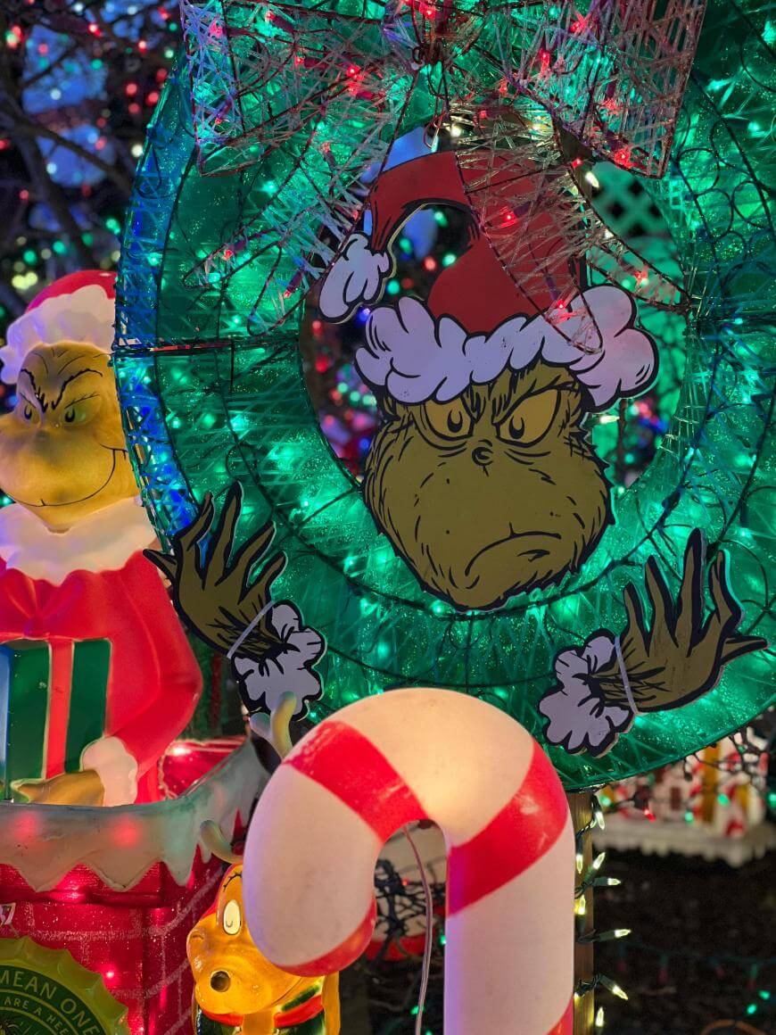 A festive holiday display featuring a large green wreath with an illustration of the Grinch wearing a Santa hat, surrounded by colorful Christmas lights, a candy cane, and a Grinch figure in a Santa outfit.