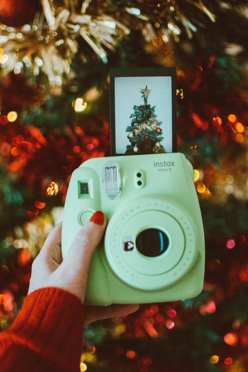 A hand holds a green Instax Mini 9 instant camera in front of a decorated Christmas tree. A photo of the tree is coming out of the camera. The person wears a red sweater and has red nail polish.