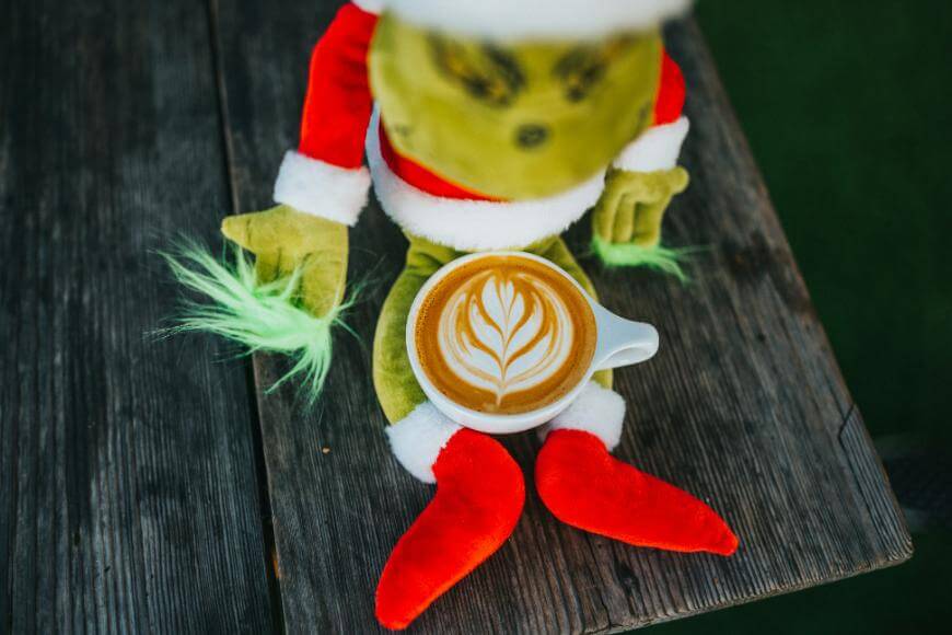 A plush Grinch doll in a Santa suit sits on a wooden surface with a cup of latte featuring leaf latte art placed in its lap.