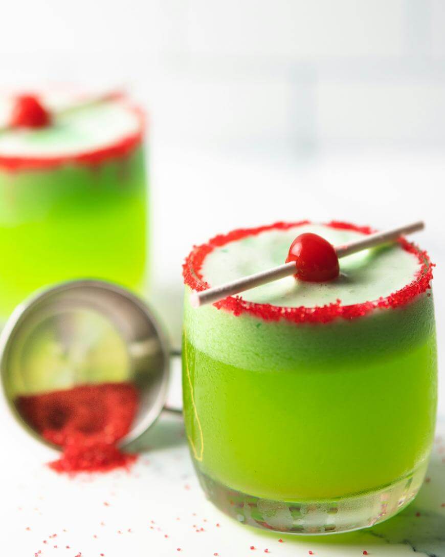A bright green cocktail in a glass with red sugar on the rim, garnished with a cherry and a stick, sits on a white surface. Another similar drink and a spilled metal shaker with red sugar are in the background.