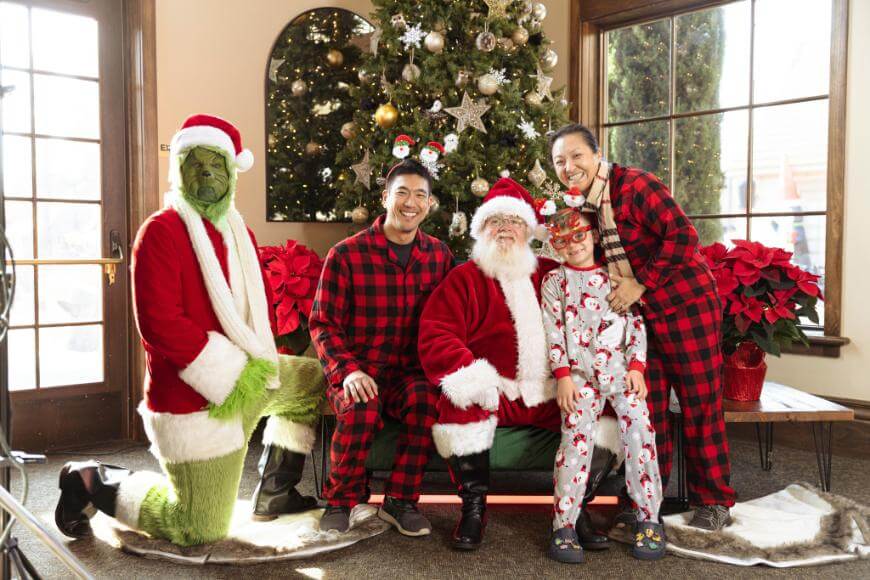 A family poses for a holiday photo with Santa Claus and the Grinch in front of a decorated Christmas tree. The family wears matching plaid pajamas, and a child sits on Santa’s lap in festive pajamas.