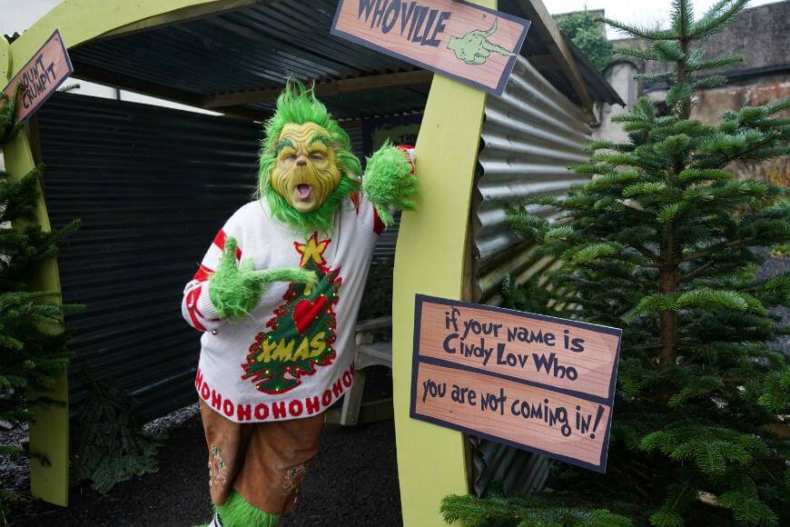 A person dressed as the Grinch, wearing a Christmas sweater, stands at an outdoor shelter with signs reading WHOVILLE and If your name is Cindy Lou Who, you are not coming in! surrounded by pine trees.