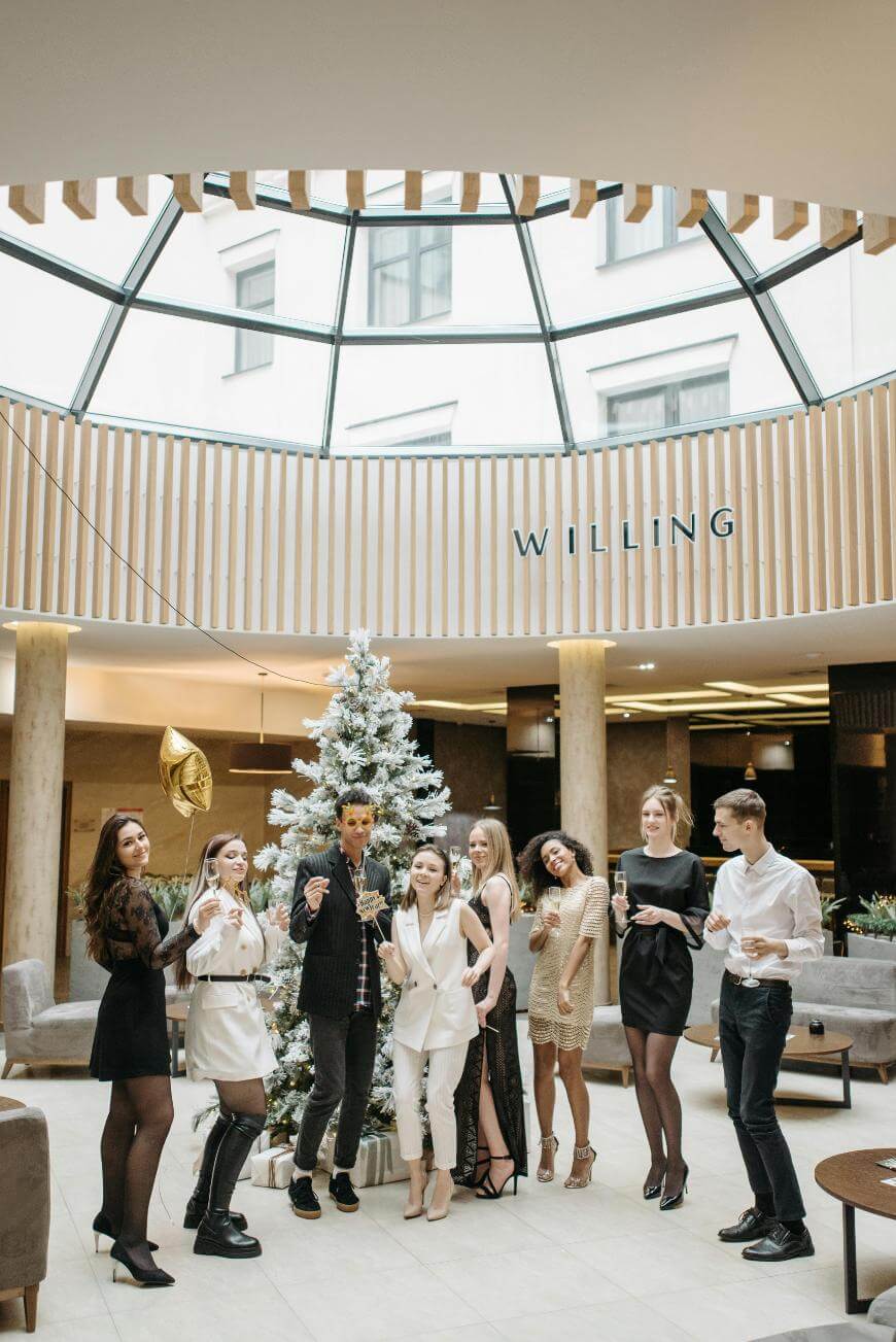 Eight people dressed in festive attire stand around a decorated Christmas tree in a modern, bright lobby, holding drinks and smiling, appearing to celebrate together. A gold balloon and the word “WILLING” are visible in the background.