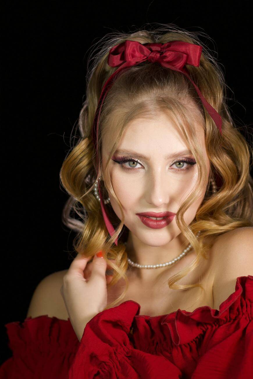 A woman with wavy blonde hair, wearing a red bow headband, off-shoulder red dress, pearl necklace, and bold makeup poses against a black background, gazing confidently at the camera.