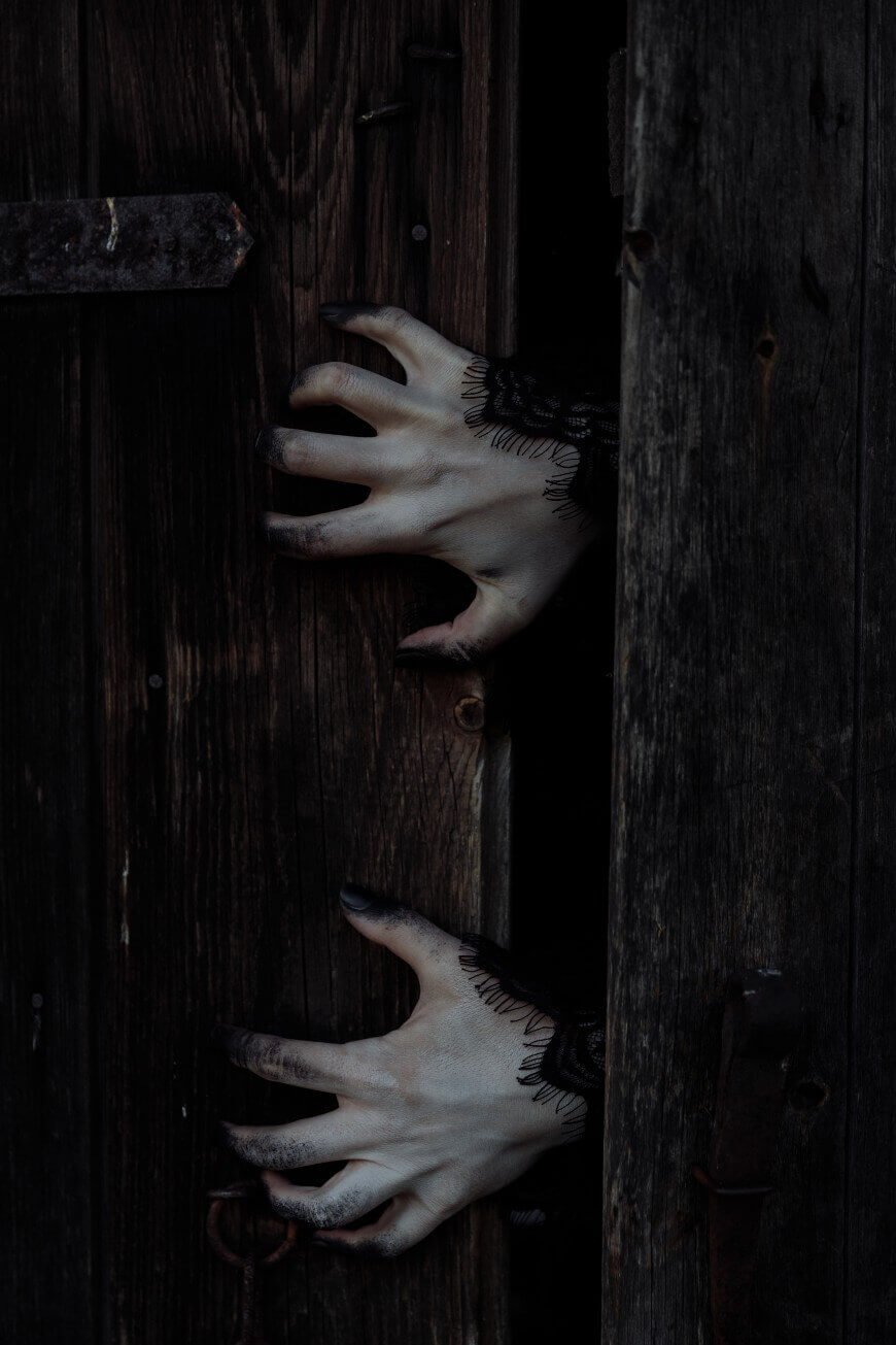 Two pale hands with black fingernails and lace cuffs reach through a narrow gap between old, dark wooden doors, creating an eerie and mysterious atmosphere.