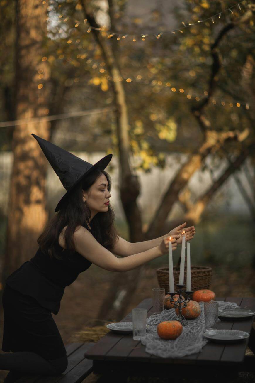 A woman dressed as a witch in a black outfit and pointed hat lights candles on an outdoor table decorated with pumpkins, plates, and glasses, in a wooded setting with string lights in the background.