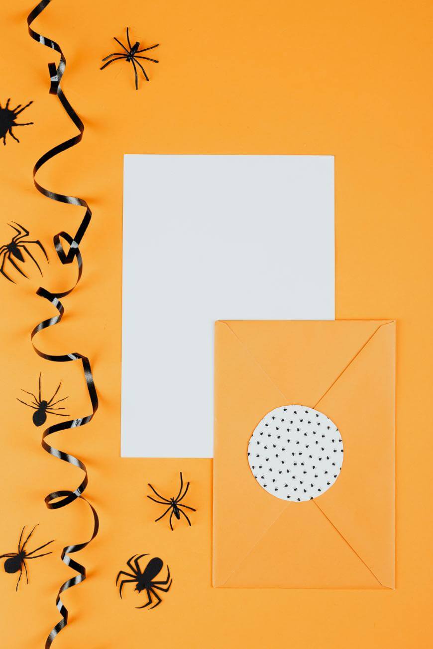 A blank white sheet of paper and an orange envelope with a polka dot seal are on an orange background, surrounded by black plastic spiders and a curled black-and-white ribbon.