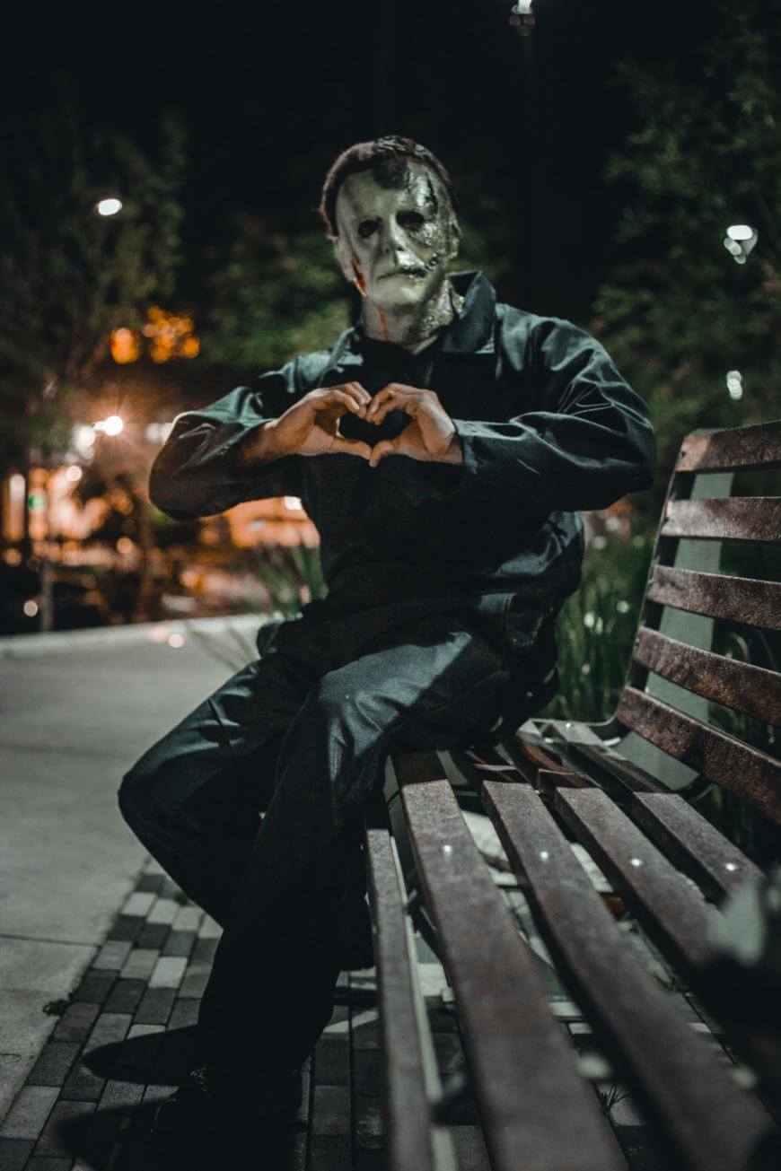 A person in a scary white mask and dark clothing sits on a wooden bench at night, making a heart shape with their hands. The background is dimly lit with trees and streetlights.
