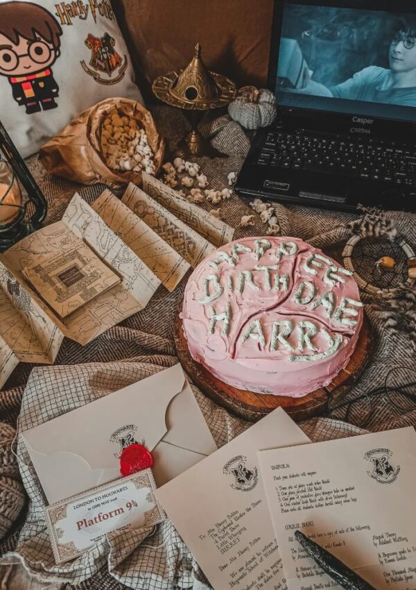 A Harry Potter-themed scene with a pink cake reading “HAPPEE BIRTHDAE HARRY,” envelopes, a Marauder’s Map, a laptop showing Harry Potter, popcorn, a lantern, and themed decorations on a cozy blanket.