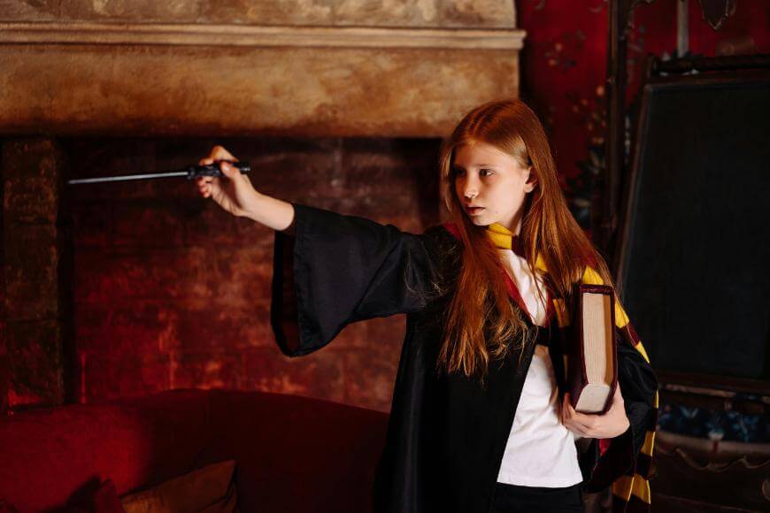 A young girl with long red hair wearing a black robe and a yellow-and-burgundy striped scarf holds a wand outstretched in one hand and a large book in the other, standing indoors against a warm, rustic background.