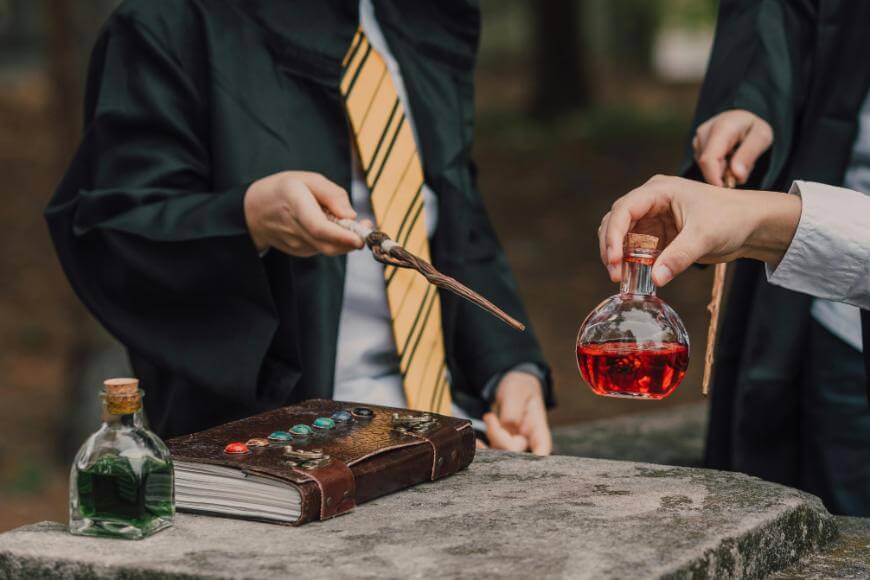 Two people in black cloaks, one with a yellow-striped tie, hold wands over a stone surface with a decorated book and potion bottles; one hand holds a round bottle filled with red liquid.