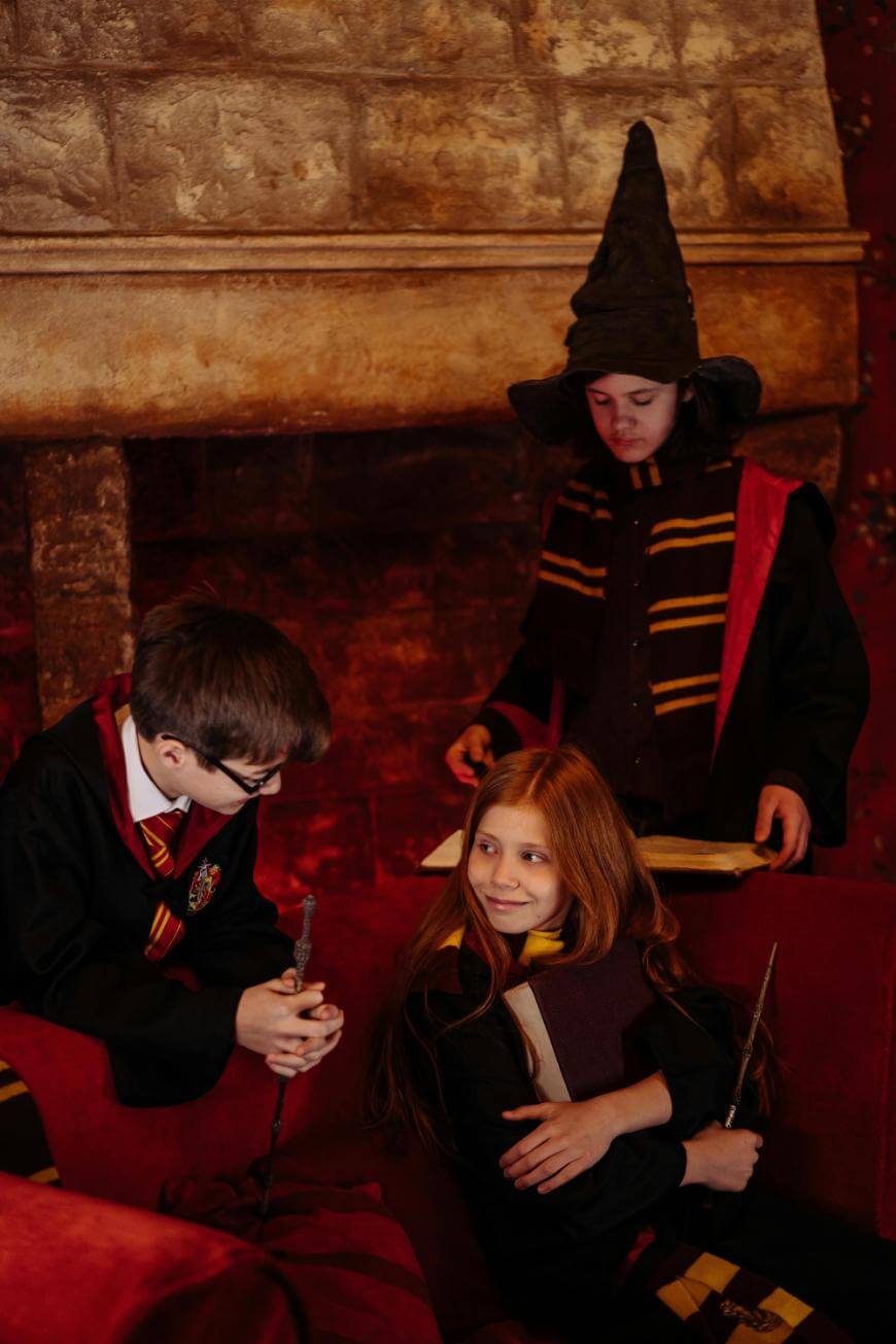 Three children dressed in wizard costumes, holding wands and wearing scarves, sit and stand in a cozy, dimly lit room with stone walls and a fireplace, resembling a magical or Harry Potter-inspired setting.