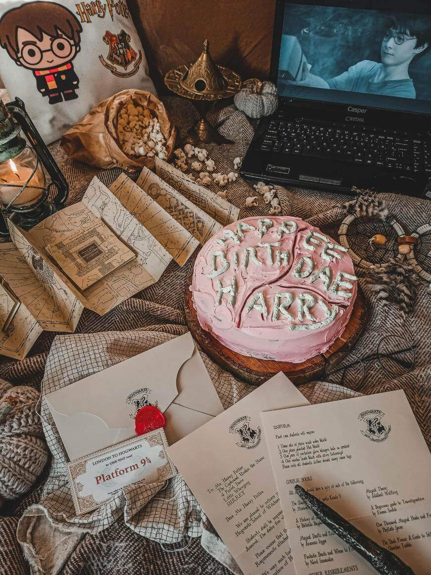 A Harry Potter-themed setting with a pink Happee Birthdae Harry cake, Hogwarts letters, a laptop playing a Harry Potter film, a lantern, a pillow, popcorn, and various magical props arranged on a cozy blanket.
