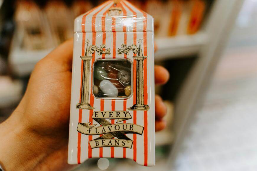 A hand holds a box of “Every Flavour Beans” with red and white stripes and a small window showing colorful jelly beans inside. The box resembles packaging from the Harry Potter series.