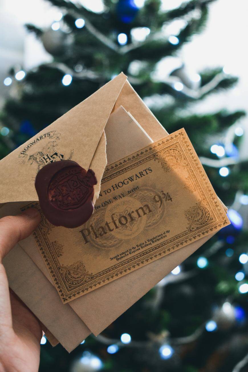 A hand holds a Hogwarts acceptance letter with a red wax seal and a golden Platform 9¾ ticket, with a blurred, decorated Christmas tree in the background.